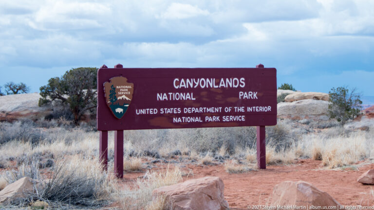 A maroon Canyonlands National Park sign stands among pale grasses and rocks beneath a cloudy sky.