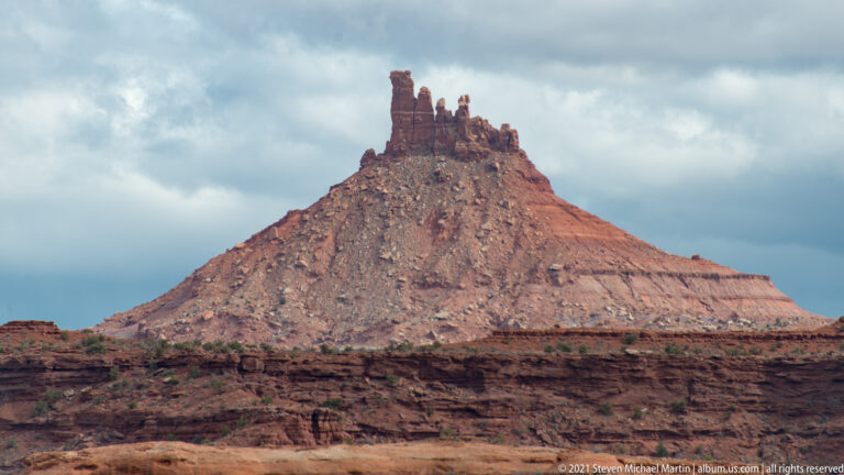 A steep red butte with a jagged pinnacled summit rises above a flat sandstone bench.