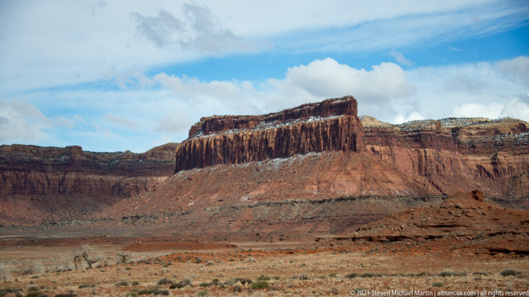 A long mesa with grooved sandstone cliffs rises above a dry, sparsely vegetated basin.