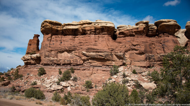 A layered red sandstone wall topped with rounded blocks rises above rubble and scattered shrubs.
