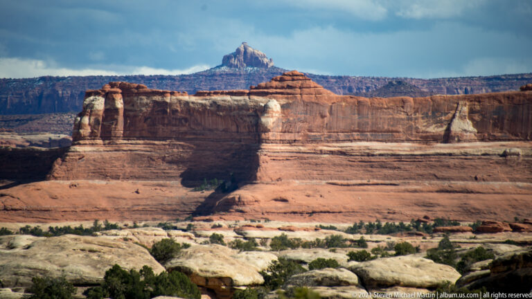 Pale bedrock overlooks a broad layered canyon wall and a distant summit beneath clouds.