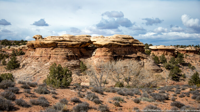 Stacked sandstone slabs form a low overhang above scrub and broken rock beneath a cloudy sky.