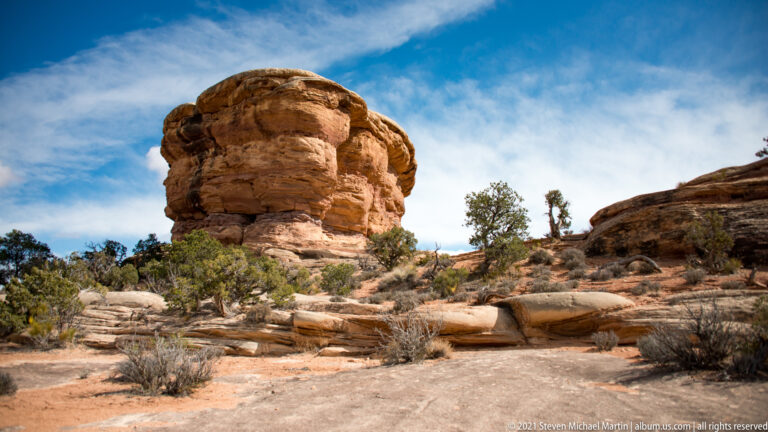 A rounded sandstone tower rises above pale slickrock and low shrubs under a blue sky.