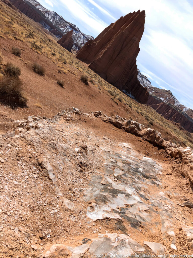 A slanted red sandstone fin rises above a gravel slope, with snow-covered ridges behind.
