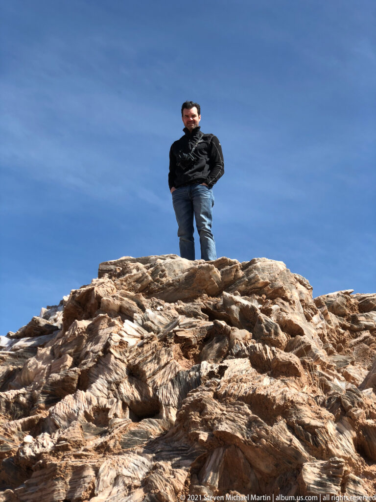 A visitor stands on a pale fractured rock crest beneath an expansive blue sky.