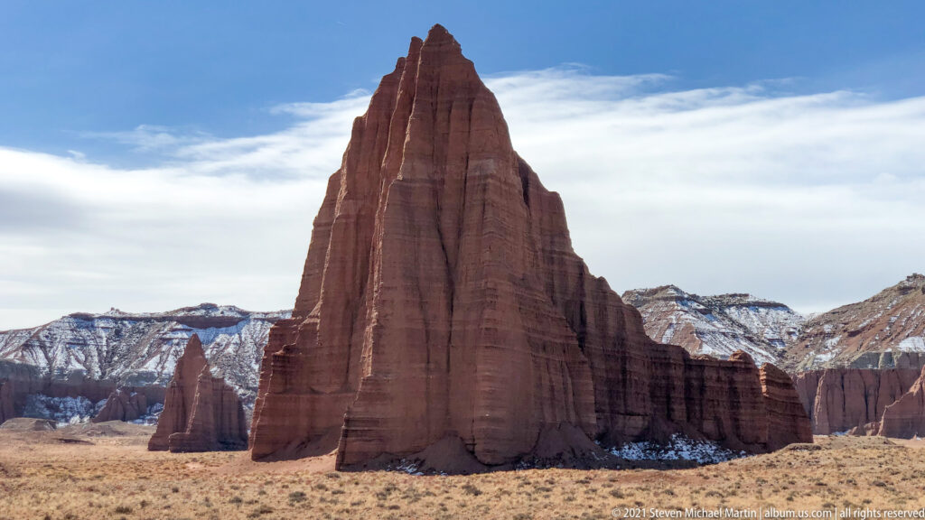 A tall red monolith rises above dry grass, with smaller spires and snow-patched hills behind.