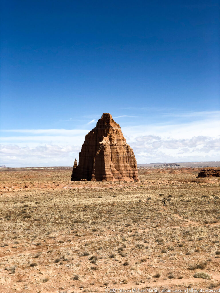 A solitary red sandstone butte stands in a broad grass-covered plain under blue sky.