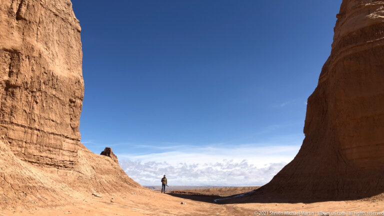 A lone hiker stands in a wide gap between tall red sandstone walls.
