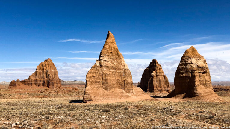A cluster of sandstone pinnacles stands beyond dry grassland and a faint curving track.