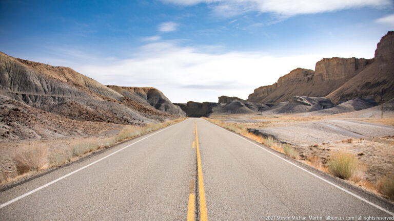 A paved two-lane road leads between steep gray-brown eroded hills beneath a lightly clouded blue sky.