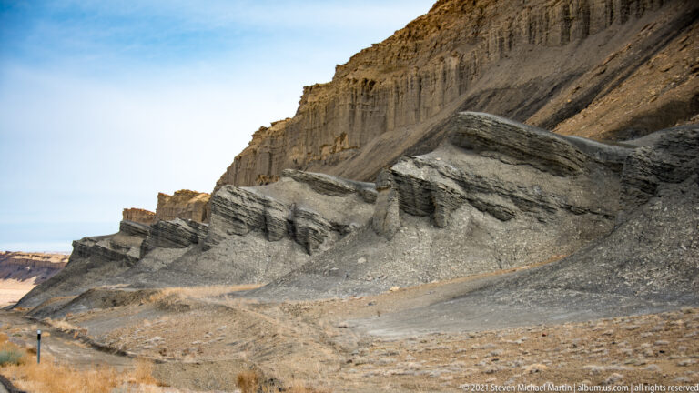 Parallel erosion grooves cut down a steep gray and tan sedimentary slope.