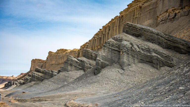 A ribbed gray-brown badlands ridge rises diagonally beneath a small patch of blue sky.