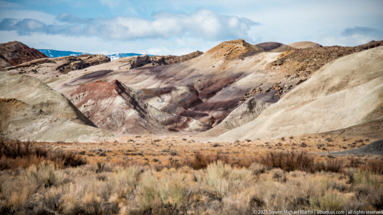 Cream, mauve, and red rounded hills rise above dry brush beneath a cloudy sky.