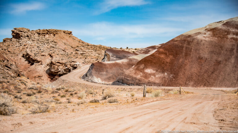 A dirt road curves between reddish and pale-banded hills, with a rough mesa at left.