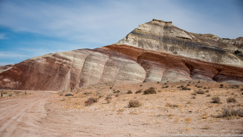 Cream, red, and gray sedimentary bands sweep across a hillside above a dirt road and scrub.