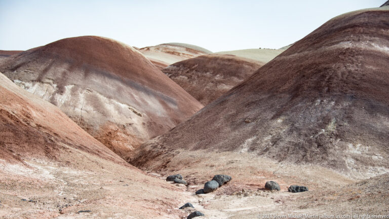 Mauve, gray, and red hillsides meet around a narrow central ravine with loose stones.