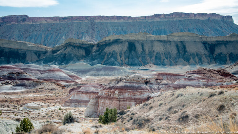 Pale and purple-banded hills stretch toward a tall dark escarpment beneath a bright sky.