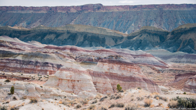 Rounded red and cream hills lie below a continuous dark cliff wall, with sparse shrubs in front.
