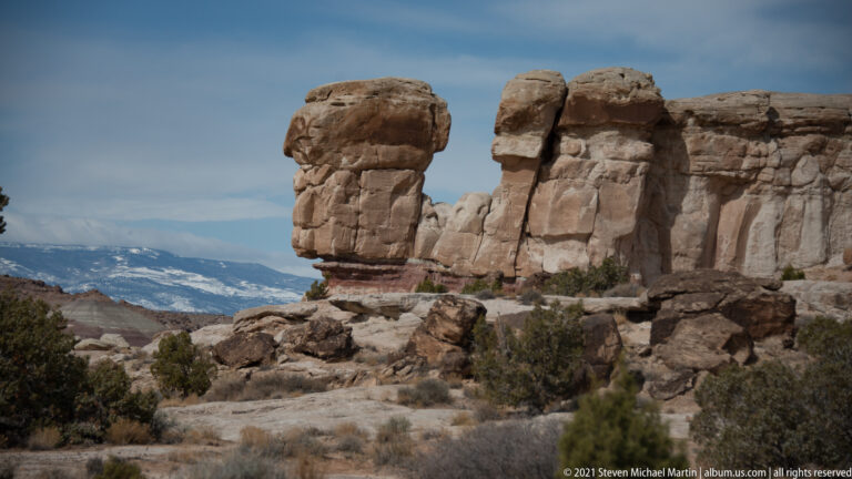 Banded cream sandstone towers stand above scrubland, with a snow-dusted mountain range stretching across the background.