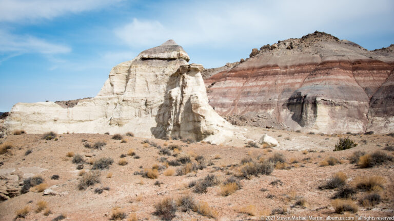 A pale eroded sandstone pillar stands before a larger cliff striped in red, gray, and cream.