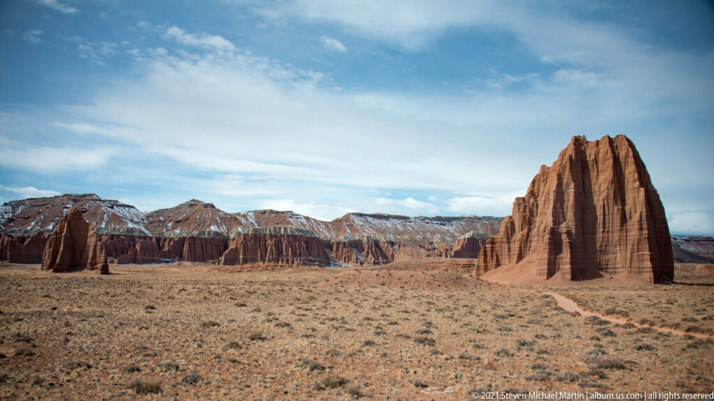 A large fluted red monolith stands at right across an open scrub plain, with a smaller tower and distant cliffs.
