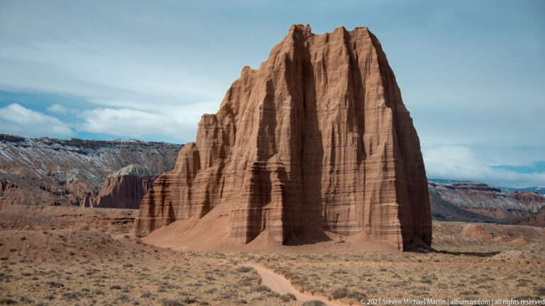 A red fluted sandstone tower rises beyond a pale dirt track crossing low vegetation.