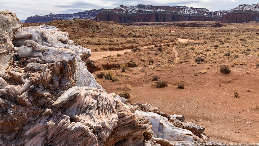 A pale veined rock outcrop overlooks red valley ground and distant dark cliffs edged with snow.