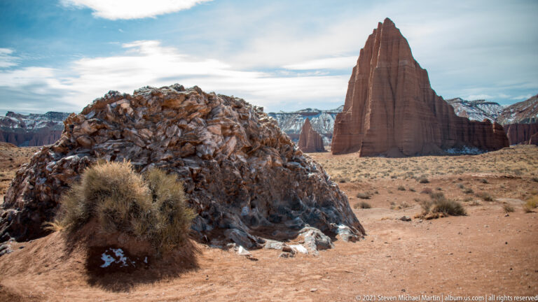 A rocky mound and dry shrub sit below a red tower, with a faint snow-dusted ridge in the distance.