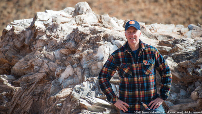 A man in a cap and plaid shirt poses before white and rust-colored layered boulders.