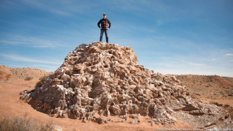 A lone person stands on the crest of a pale rocky mound against a wide blue sky.