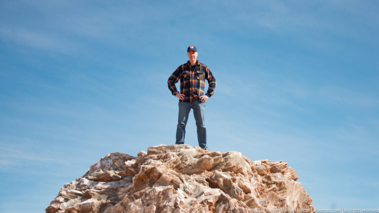 From a low angle, a man stands atop a pale boulder beneath a large expanse of blue sky.