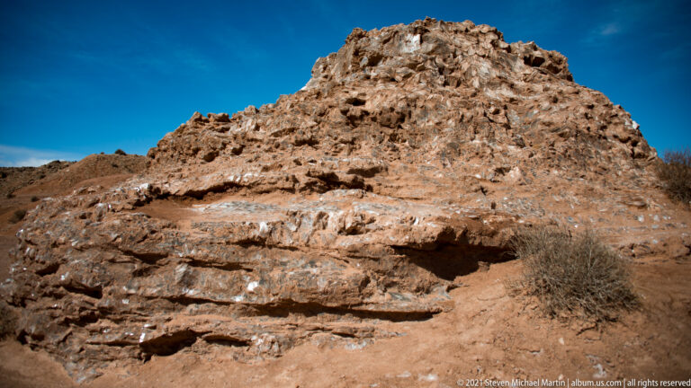 A coarse pale rock mound has a shallow recess, with a dry shrub at its right edge.