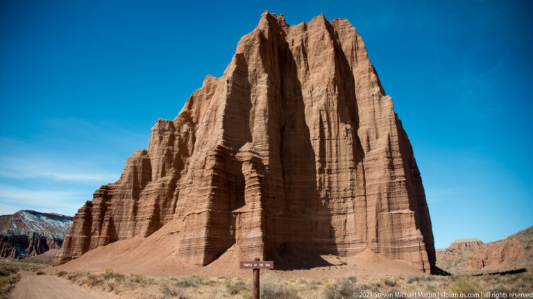 A red fluted sandstone monolith rises above scrub, with a small roadside sign near its base.