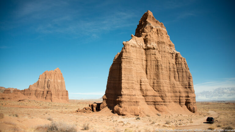 Two red sandstone towers stand beyond a dirt road, with a dark vehicle in the foreground.