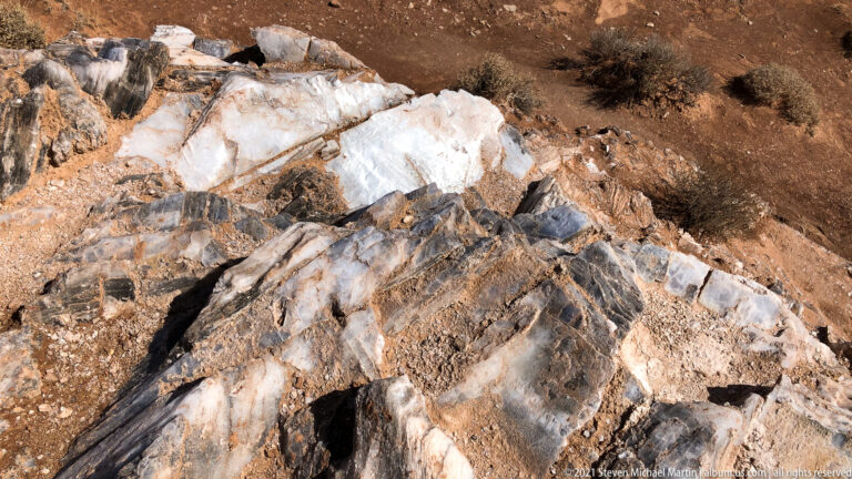 Close view of white and gray bands crossing fractured rust-colored rock and granular soil.