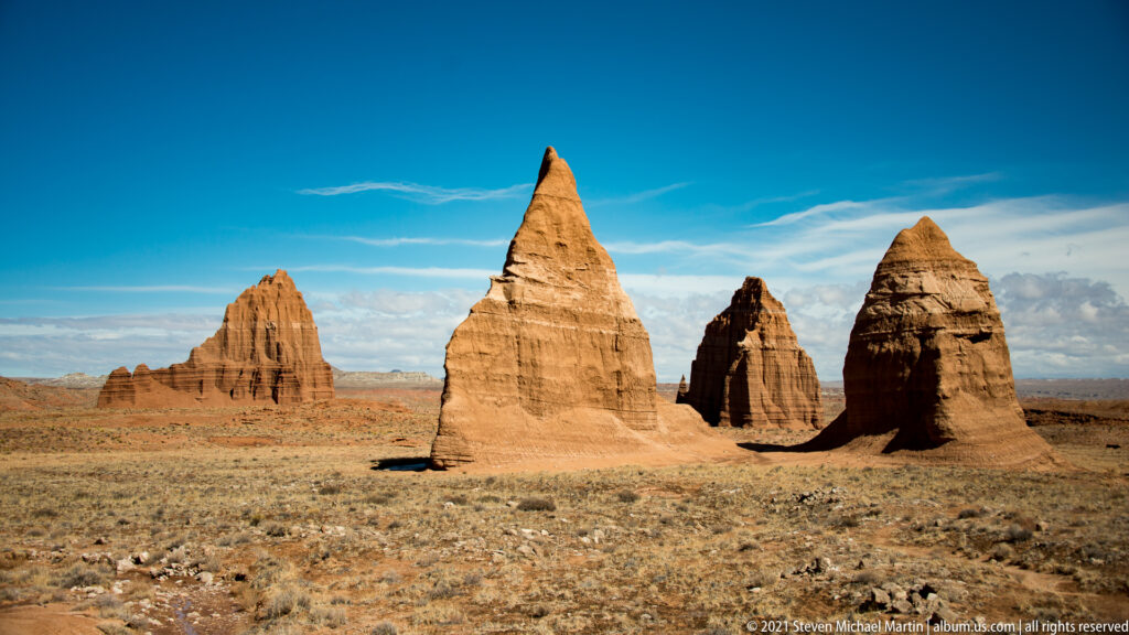 Several sandstone monoliths are spaced across a broad plain beneath a blue sky with scattered clouds.