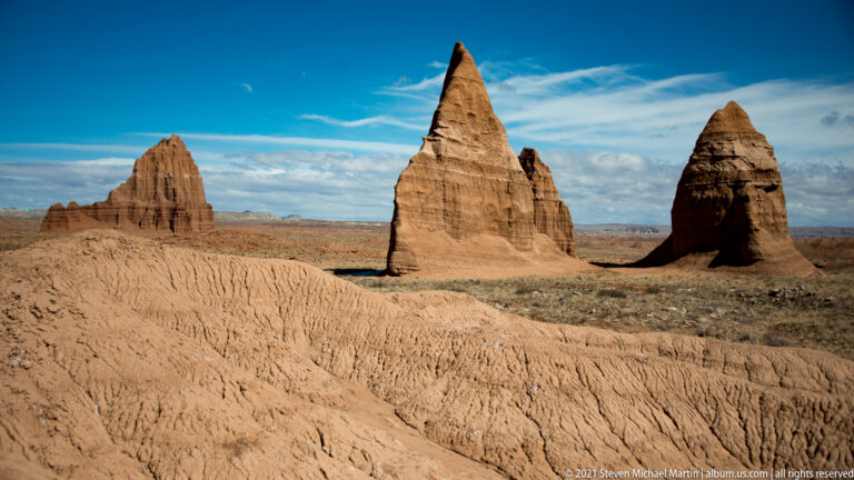A shallowly eroded slope fills the foreground, with three distant sandstone towers beneath the sky.