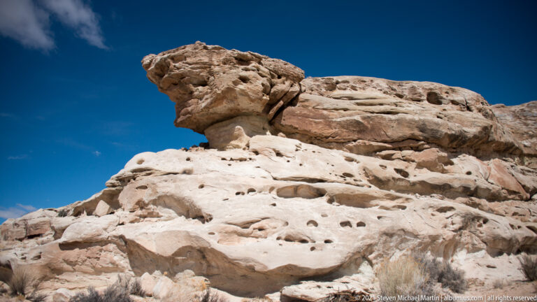 A small cap-shaped rock stands on a pale, pitted ledge beneath open sky.