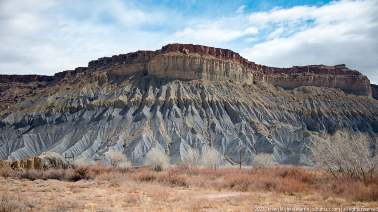 Blue-gray gullies descend beneath a dark flat-topped mesa, with grasses and shrubs in the foreground.