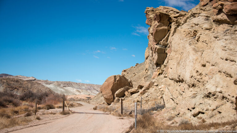A dirt road curves beside a tall pale rock wall, marked by fence posts beneath blue sky.