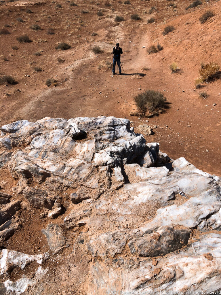 A visitor with a camera and tripod stands on red ground beyond a pale fractured rock outcrop.