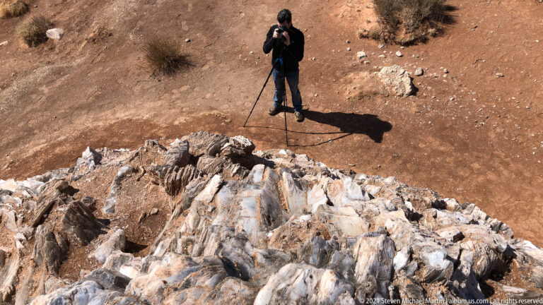 A photographer with a tripod stands above a pale fractured outcrop on reddish ground.