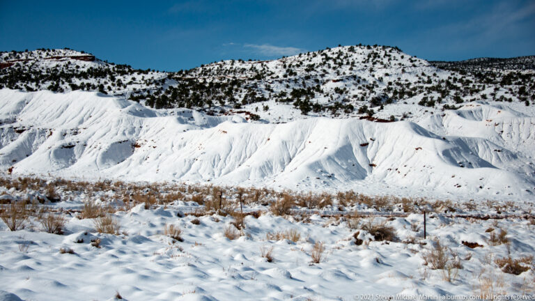 Snow-covered slopes and scattered dark shrubs extend to a low ridge beneath blue sky.