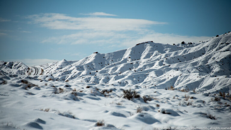 A paved road curves between snow-covered, deeply eroded slopes and distant scrubby ridges.