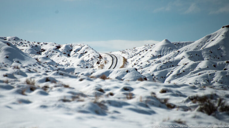 A narrow road winds between snow-covered hills, with blurred snow and scrub in the foreground.