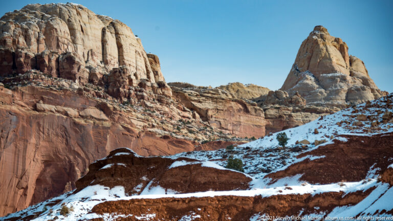A pale pointed butte rises among red cliffs with snow on ledges and slopes.