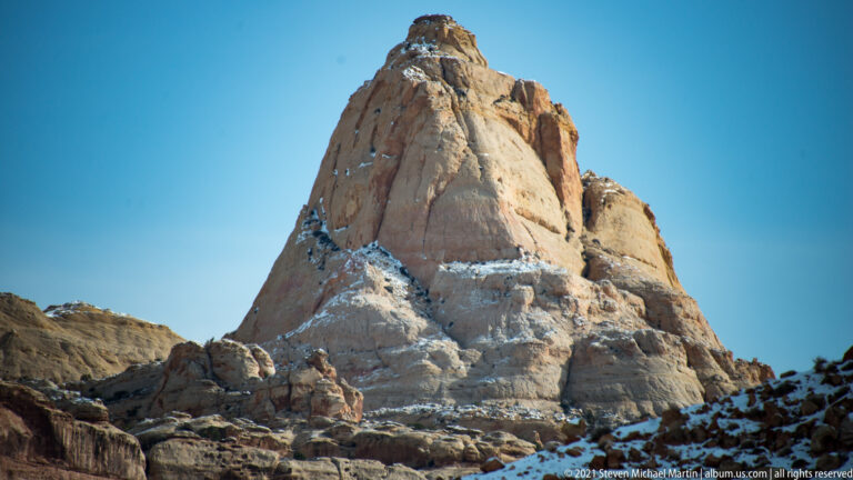 A rounded pale butte rises above layered ledges, with small snow patches on its face.