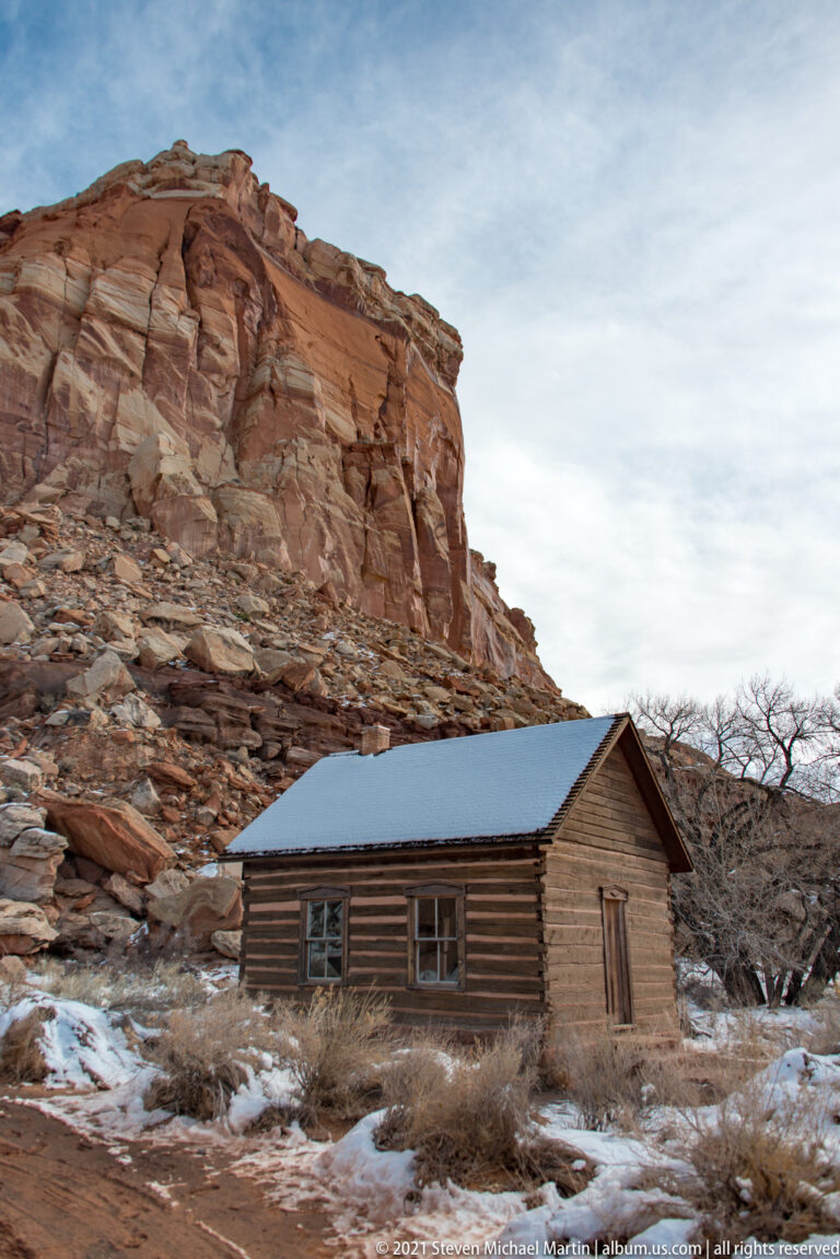 A log cabin with a snowy roof sits below a steep red-rock slope among bare trees.