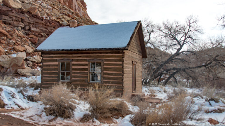 A snow-covered gabled roof sits above log walls, two windows, and a side door.