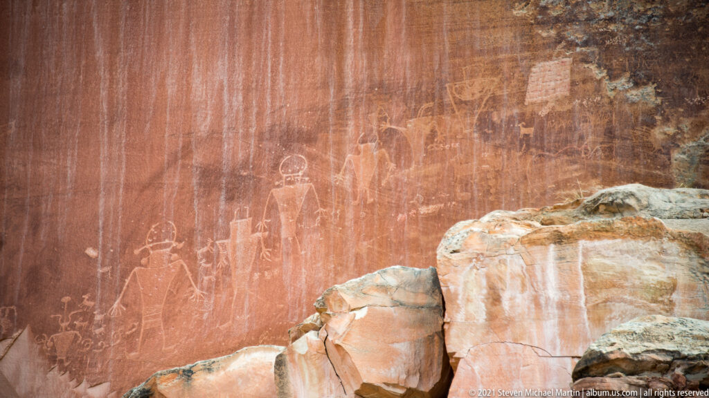 Faded pale figures and symbols cover a red sandstone panel above a rocky ledge.